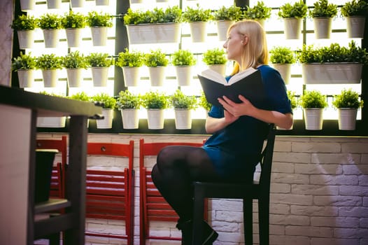 blonde woman in a dress sits on a chair with a book in hand, against the backdrop of houseplants. engaged in reading