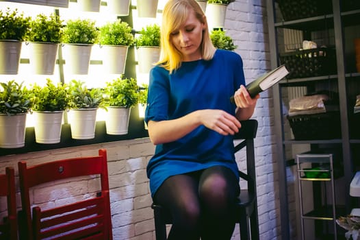 blonde woman in a dress sits on a chair with a book in hand, against the backdrop of houseplants. engaged in reading