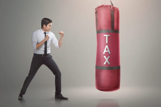 Handsome asian businessman ready to punch boxing bag with tax text on it. Taxation concept