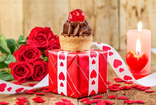 Cupcake with cherry in front of bouquet of red roses and candles on wooden background. Valentines day concept. Focus on cake.