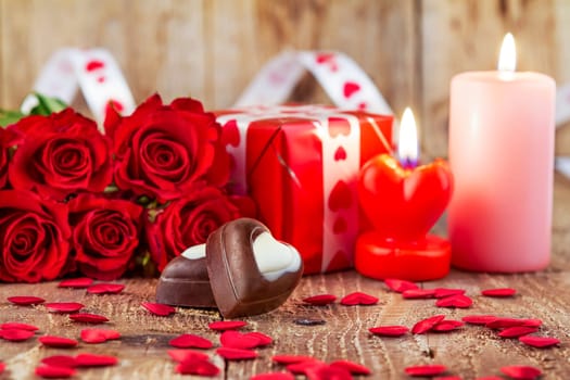 Chocolate pralines in front of bouquet of red roses and candles on wooden background. Valentines day concept. Mother’s day concept. Focus on pralines.