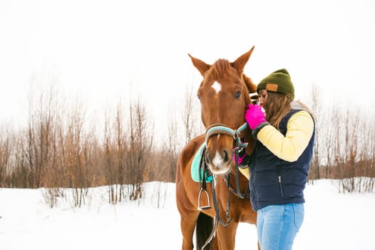 Woman in winter clothes and the horse to stand together against the white snow. love and care for horses. girl puts a bridle on a horse