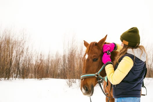 Woman in winter clothes and the horse to stand together against the white snow. love and care for horses. girl puts a bridle on a horse