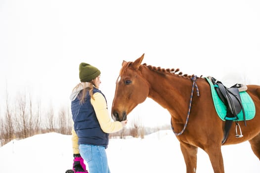 Woman in winter clothes and the horse stand together against the white snow. love and care for horses. animal portrait on a white background.