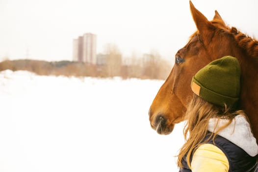 Woman in winter clothes and the horse stand together against the white snow, looking into the distance in the city homes. love and care for horses