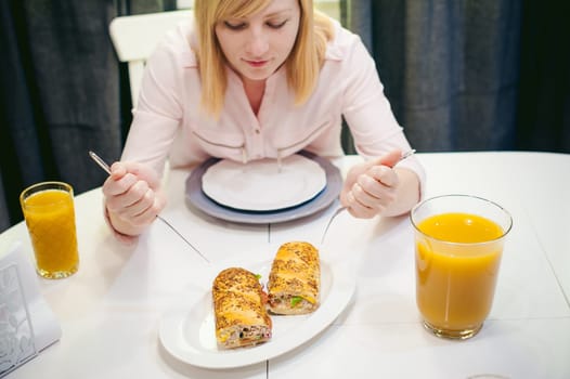 Pretty blonde woman in a pink shirt, eating at home a fork and knife. breakfast sandwich and peach juice. on white table in bright interior of kitchen in plate is sandwich juice poured in glass