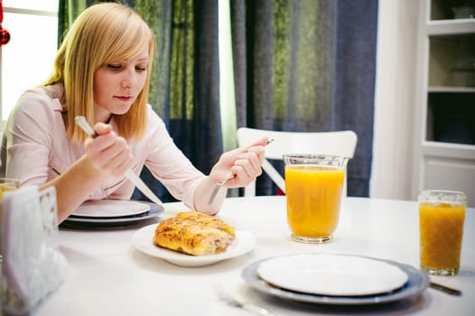 Pretty blonde woman in a pink shirt, eating at home a fork and knife. breakfast sandwich and peach juice. on white table in bright interior of kitchen in plate is sandwich juice poured in glass