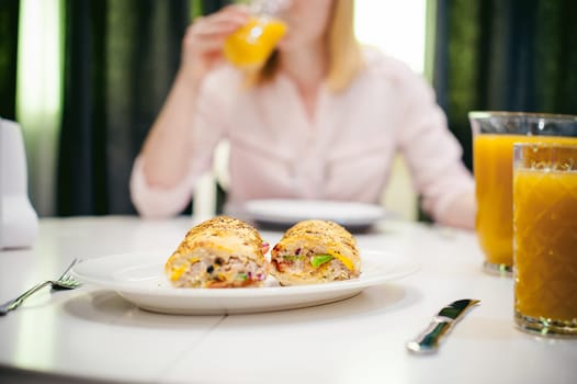 Pretty blonde woman in a pink shirt, eating at home. breakfast sandwich and peach juice. on white table in bright interior of kitchen in plate is sandwich juice poured in glass