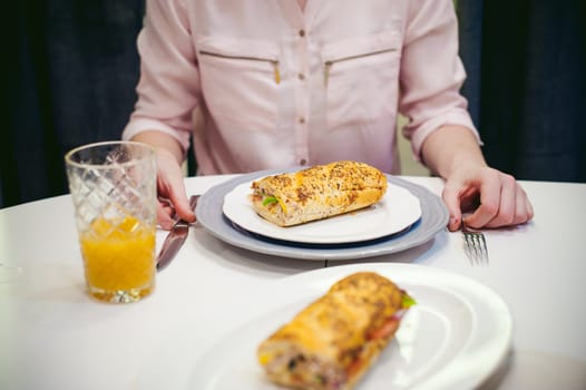 Pretty blonde woman in a pink shirt, eating at home. breakfast sandwich and peach juice. on white table in bright interior of kitchen in plate is sandwich juice poured in glass