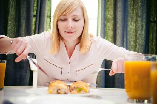 Pretty blonde woman in a pink shirt, eating at home a fork and knife. breakfast sandwich and peach juice. on white table in bright interior of kitchen in plate is sandwich juice poured in glass