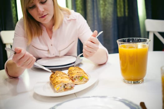Pretty blonde woman in a pink shirt, eating at home a fork and knife. breakfast sandwich and peach juice. on white table in bright interior of kitchen in plate is sandwich juice poured in glass