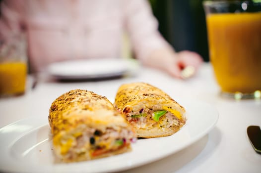Pretty blonde woman in a pink shirt, eating at home. breakfast sandwich and peach juice. on white table in bright interior of kitchen in plate is sandwich juice poured in glass