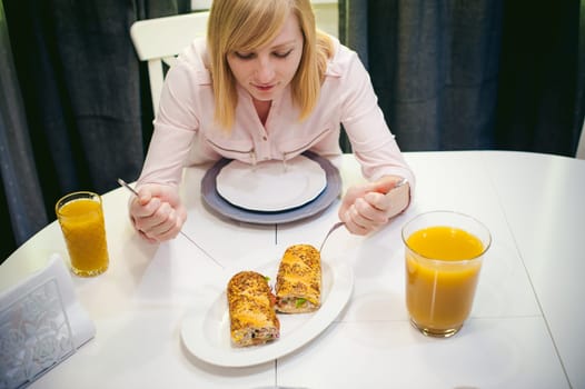 Pretty blonde woman in a pink shirt, eating at home a fork and knife. breakfast sandwich and peach juice. on white table in bright interior of kitchen in plate is sandwich juice poured in glass