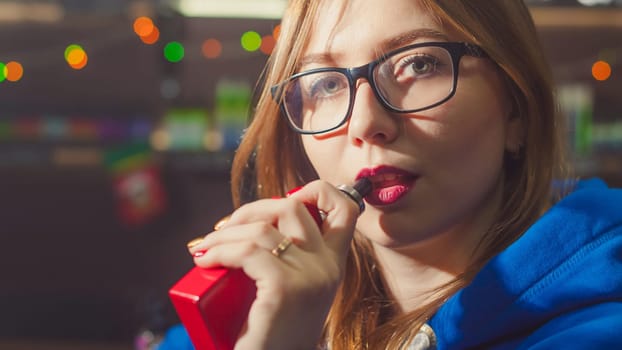 beautiful young woman with glasses smoking electronic cigarette. girl in a blue sweater brings to his lips red box mod, sitting indoors vape shop cafe