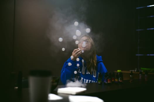 young Cute woman, the seller in vape shop shop, smoking electronic cigarette,  vaping and releases a cloud of vapor and inflates soap bubbles