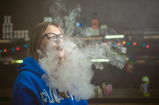 beautiful young woman with glasses smoking electronic cigarette. girl in a blue sweater blows clouds of glycerol vapor, from vape mod, sitting indoors vape shop cafe