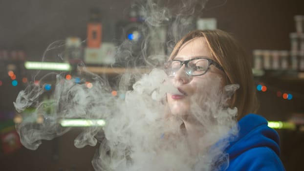 beautiful young woman with glasses smoking electronic cigarette. girl in a blue sweater blows clouds of glycerol vapor, from vape mod, sitting indoors vape shop cafe