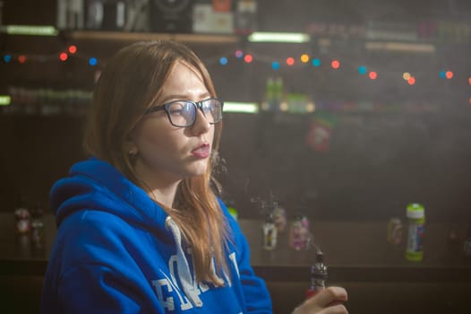 beautiful young woman with glasses smoking electronic cigarette. girl in a blue sweater blows clouds of glycerol vapor, from vape mod, sitting indoors vape shop cafe