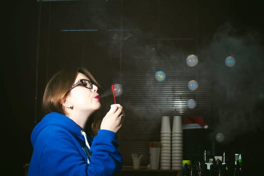 young Cute woman, the seller in vape shop shop, smoking electronic cigarette,  vaping and releases a cloud of vapor and inflates soap bubbles