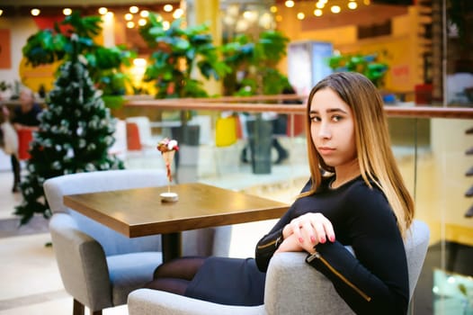 young woman in a black dress is sitting in a chair against a background of interior cafe in a shopping center