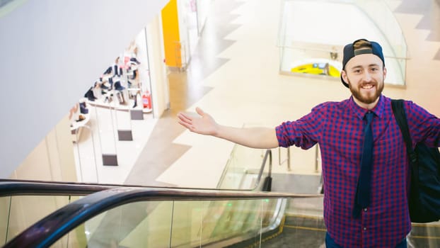 young man with a beard in a plaid shirt and a tie and cap, riding on an escalator in the mall