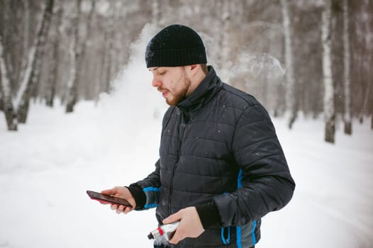 a young man with a beard, smokes an electronic cigarette and happily talking on the phone through the Internet, a positive relaxing outdoors in the snow in the winter