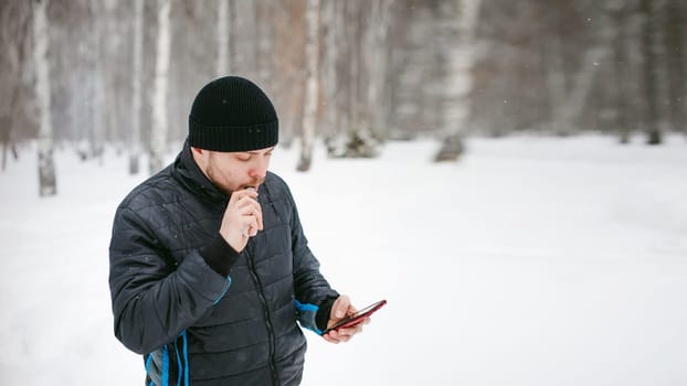 a young man with a beard, smokes an electronic cigarette and happily talking on the phone through the Internet, a positive relaxing outdoors in the snow in the winter