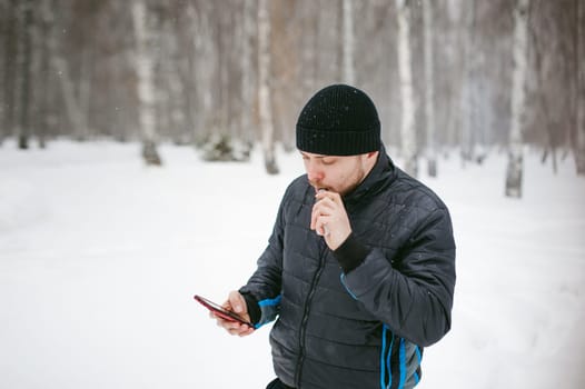 a young man with a beard, smokes an electronic cigarette and happily talking on the phone through the Internet, a positive relaxing outdoors in the snow in the winter
