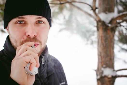 a young man with a beard, smokes an electronic cigarette, outdoors in winter snow