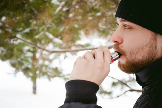 a young man with a beard, smokes an electronic cigarette, outdoors in winter snow