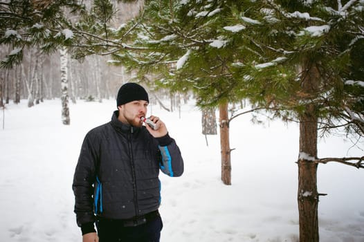 a young man with a beard, smokes an electronic cigarette, outdoors in winter snow