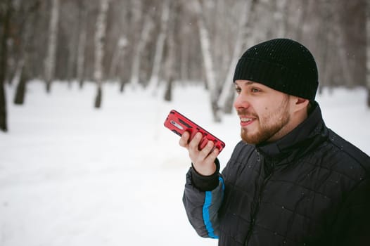a young man with a beard, smokes an electronic cigarette and happily talking on the phone through the Internet, a positive relaxing outdoors in the snow in the winter