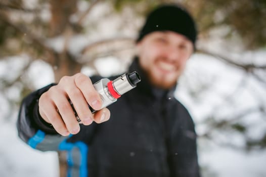 a young man with a beard, smokes an electronic cigarette, outdoors in winter snow