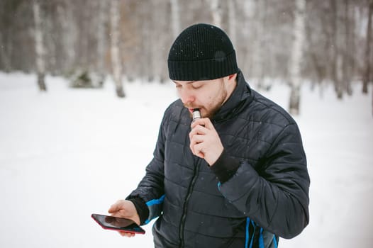 a young man with a beard, smokes an electronic cigarette and happily talking on the phone through the Internet, a positive relaxing outdoors in the snow in the winter