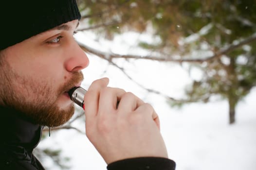 a young man with a beard, smokes an electronic cigarette, outdoors in winter snow