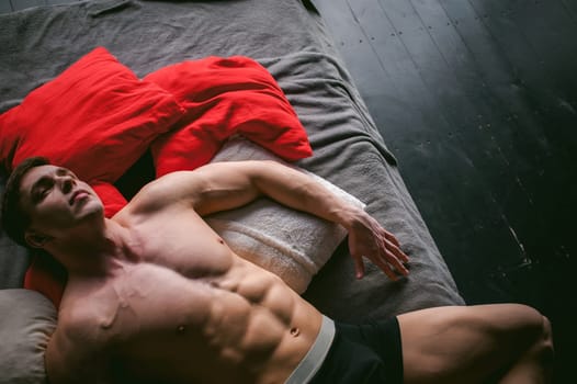 studio portrait young sexy men bodybuilder athlete, with a bare torso, lies on a bed in pillows in underwear