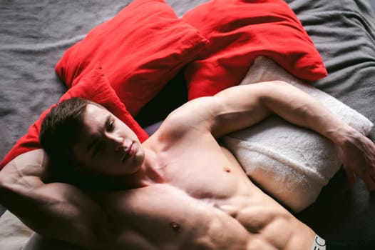 studio portrait young sexy men bodybuilder athlete, with a bare torso, lies on a bed in pillows in underwear