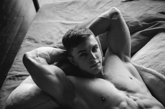 studio portrait young sexy men bodybuilder athlete, with a bare torso, lies on a bed in pillows in underwear