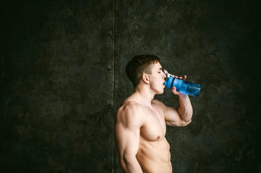 Young sexy men bodybuilder athlete,studio portrait loft on background of stylized wall,guy model With a bare torso Drinks water from a sports bottle of blue color after training