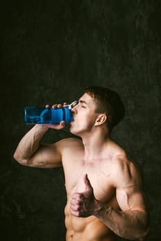 Young sexy men bodybuilder athlete,studio portrait loft on background of stylized wall,guy model With bare torso Drinks water from sports bottle of blue color after training, shows gesture thumbs up