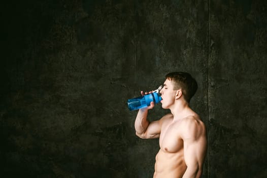 Young sexy men bodybuilder athlete,studio portrait loft on background of stylized wall,guy model With a bare torso Drinks water from a sports bottle of blue color after training