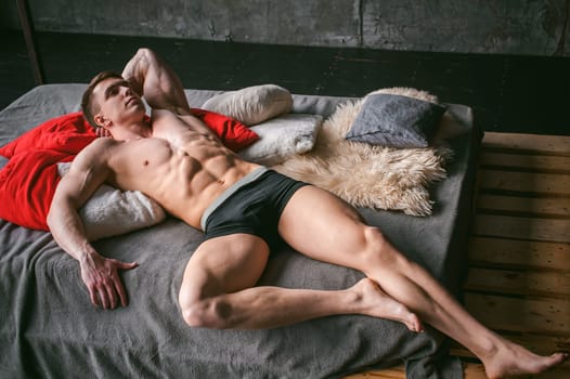 studio portrait young sexy men bodybuilder athlete, with a bare torso, lies on a bed in pillows in underwear