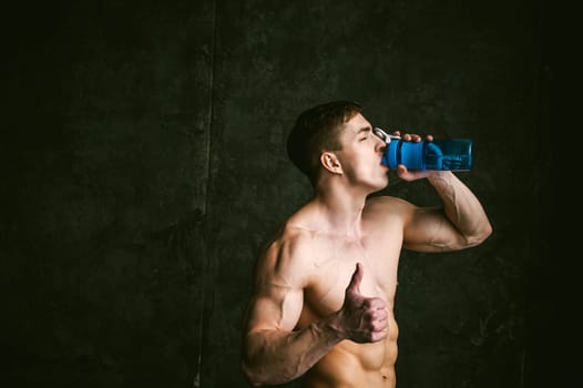 Young sexy men bodybuilder athlete,studio portrait loft on background of stylized wall,guy model With bare torso Drinks water from sports bottle of blue color after training, shows gesture thumbs up
