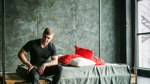 Young sexy men bodybuilder athlete,studio portrait loft on background of stylized wall,guy model black Tshirt and trousers Sits on bed with pillows and rug on brutal male bed made of metal and pallets