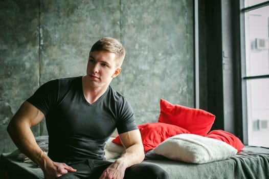 Young sexy men bodybuilder athlete,studio portrait loft on background of stylized wall,guy model black Tshirt and trousers Sits on bed with pillows and rug on brutal male bed made of metal and pallets