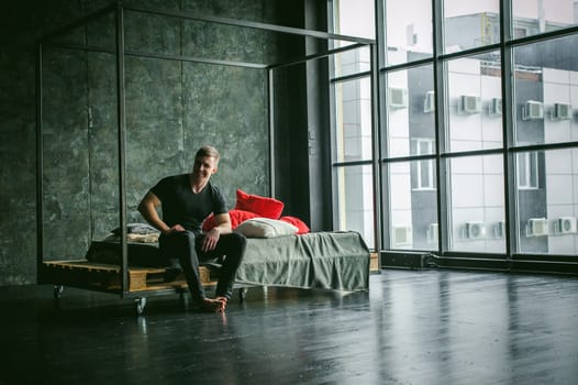 Young sexy men bodybuilder athlete,studio portrait loft on background of stylized wall,guy model black Tshirt and trousers Sits on bed with pillows and rug on brutal male bed made of metal and pallets