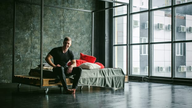 Young sexy men bodybuilder athlete,studio portrait loft on background of stylized wall,guy model black Tshirt and trousers Sits on bed with pillows and rug on brutal male bed made of metal and pallets