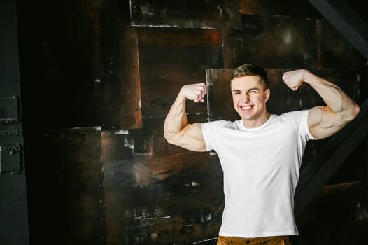Young sexy men bodybuilder athlete, studio portrait in a loft on a background of  stylized metal wall, a guy model in a white T-shirt and brown trousers, tensed his arms showing muscles, bared teeth