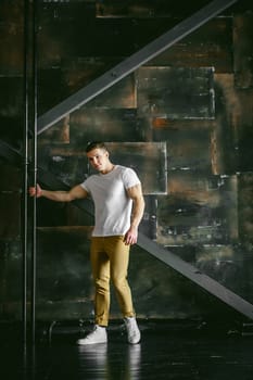 Young sexy men bodybuilder athlete, Studio portrait in a loft on the background of a stylized metal wall, a guy model in a white T-shirt and brown trousers