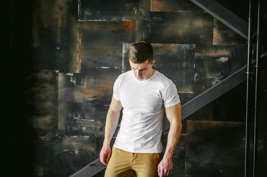 Young sexy men bodybuilder athlete, Studio portrait in a loft on the background of a stylized metal wall, a guy model in a white T-shirt and brown trousers